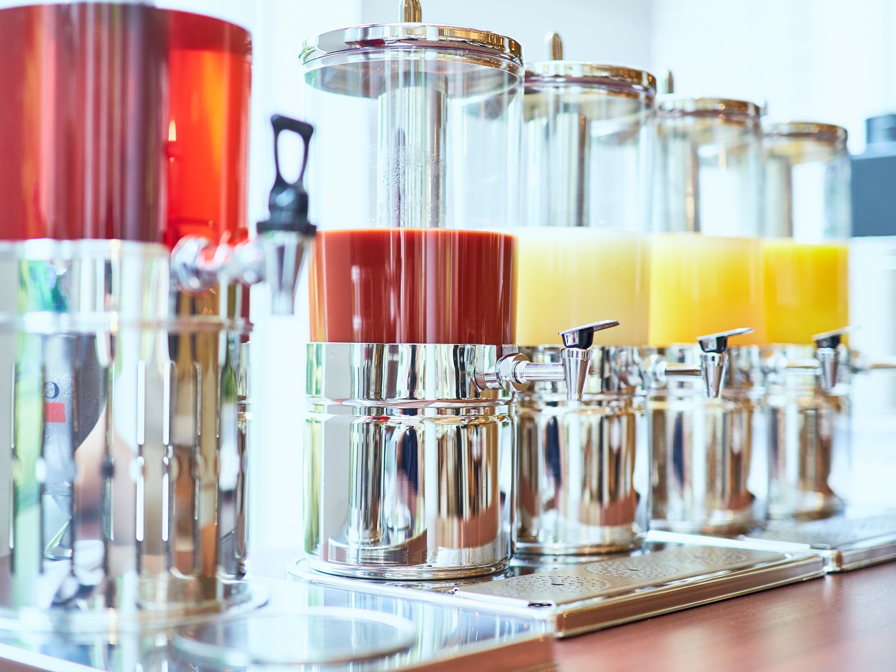 Een rij roestvrijstalen dispensers met transparante glazen deksels, vermoedelijk voor drankjes, op een reflecterend oppervlak.