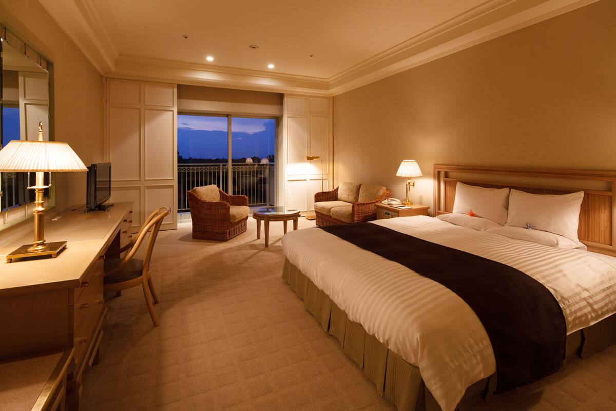 A hotel room with a large bed, two chairs, a desk with a lamp, a TV, and a balcony with a view.
