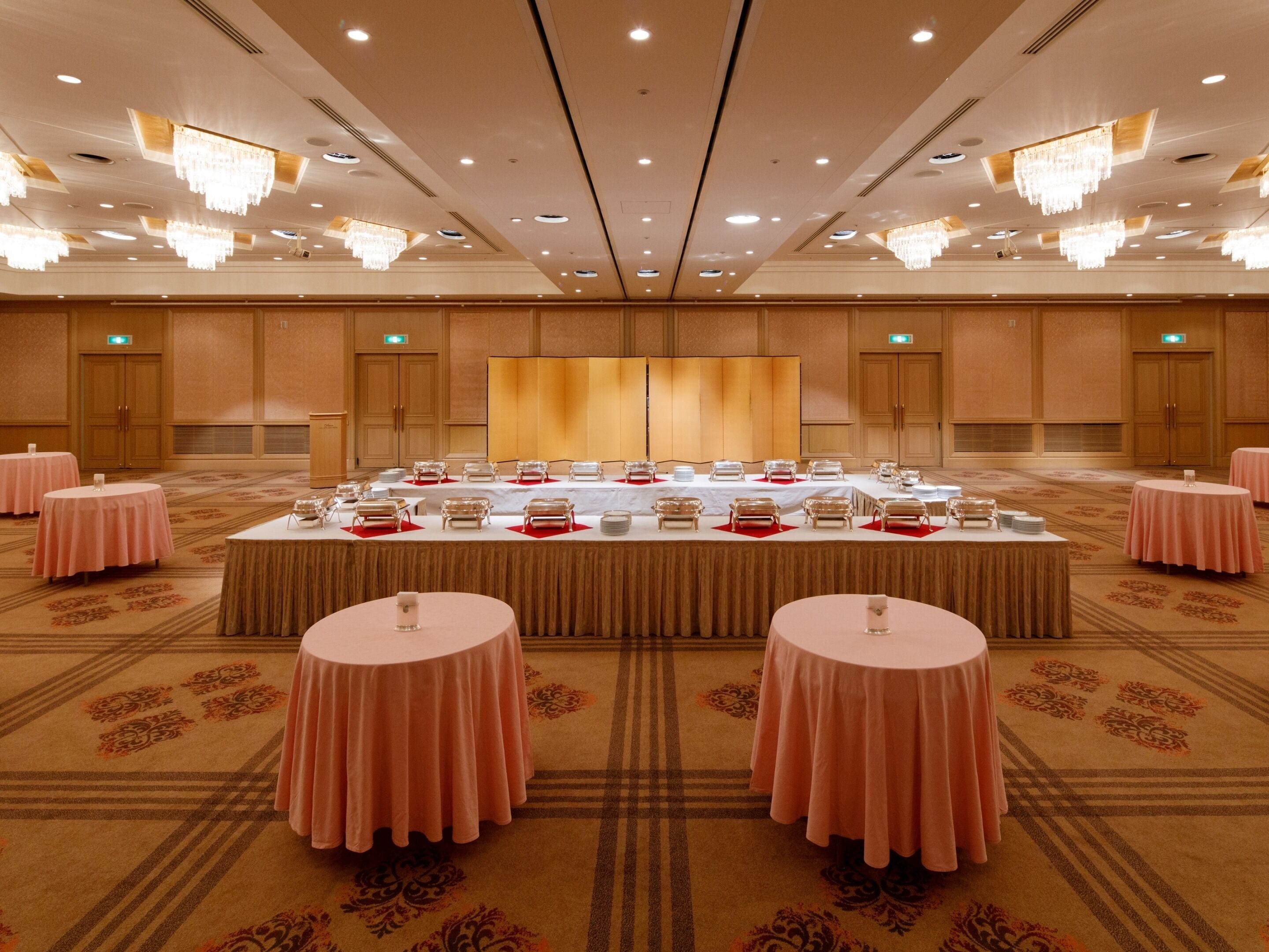 Un amplio salón de conferencias con una larga mesa preparada para una reunión, rodeada de mesas circulares más pequeñas, y un suelo alfombrado con un diseño geométrico.