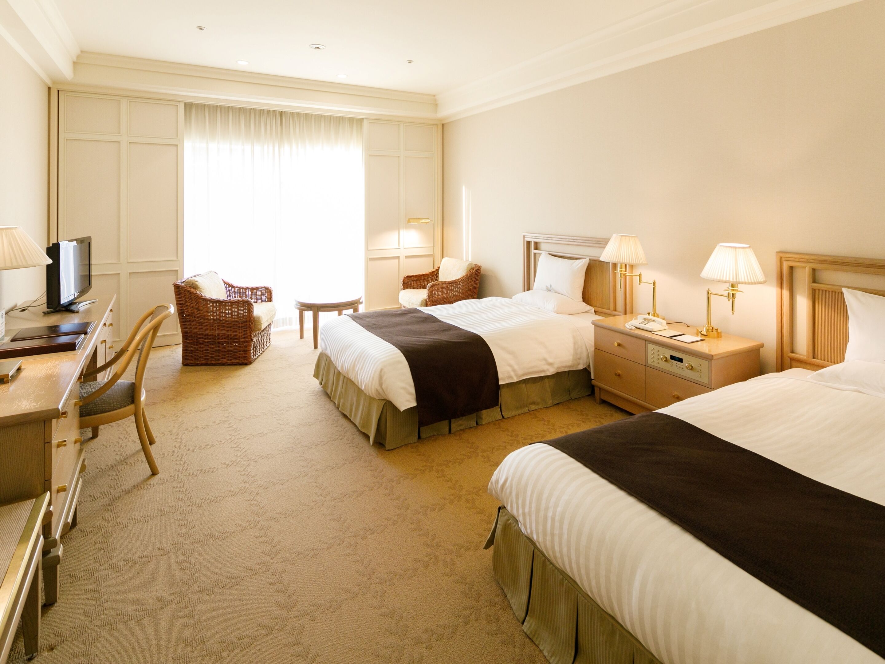 Habitación de hotel con dos camas, un escritorio con televisor, una silla, una mesita y una ventana con cortinas.