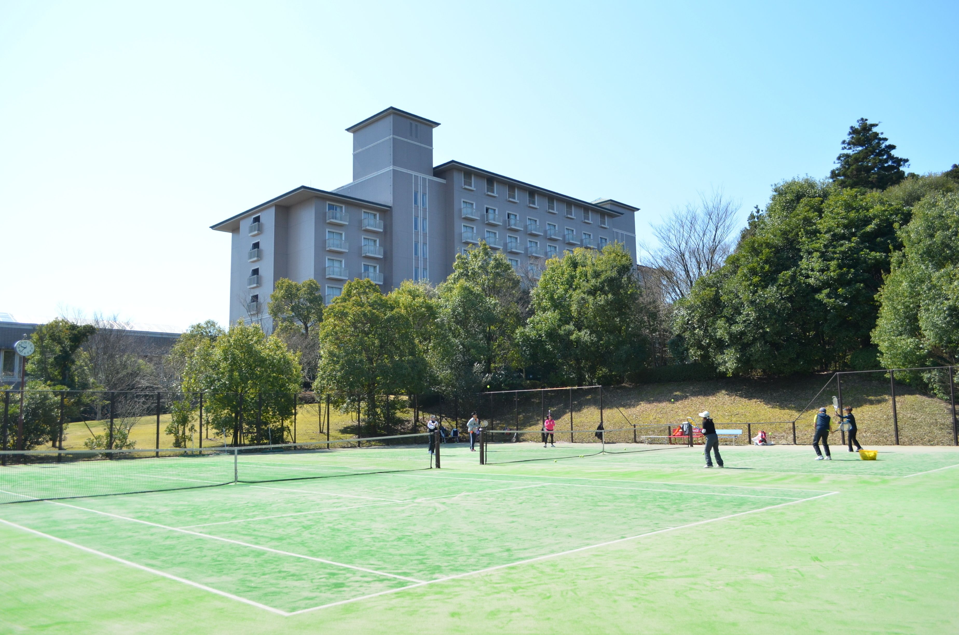 Un court de tennis avec des joueurs, entouré d’arbres et avec un bâtiment en arrière-plan.