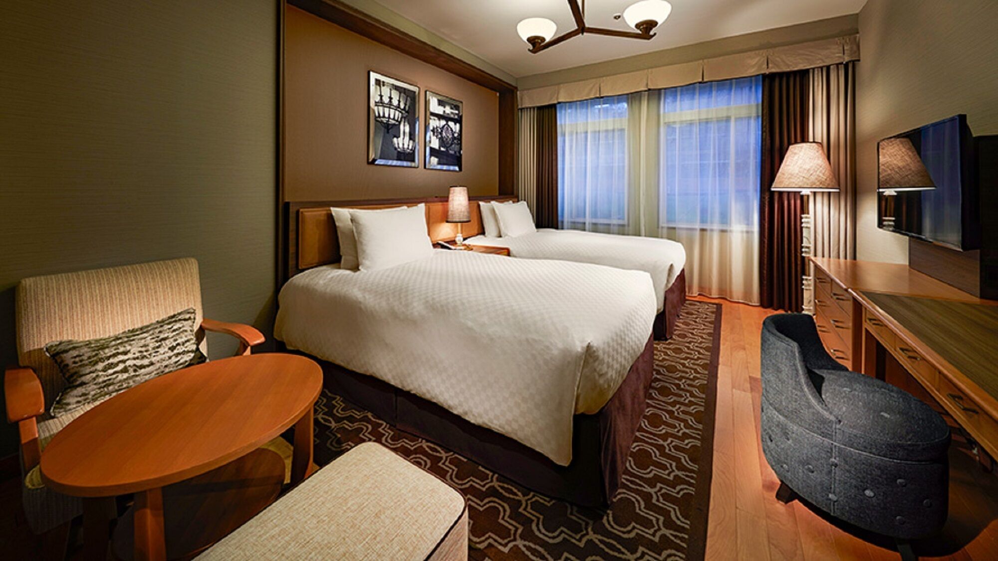 A hotel room with two beds, a desk, a chair, a TV, and a window with curtains.