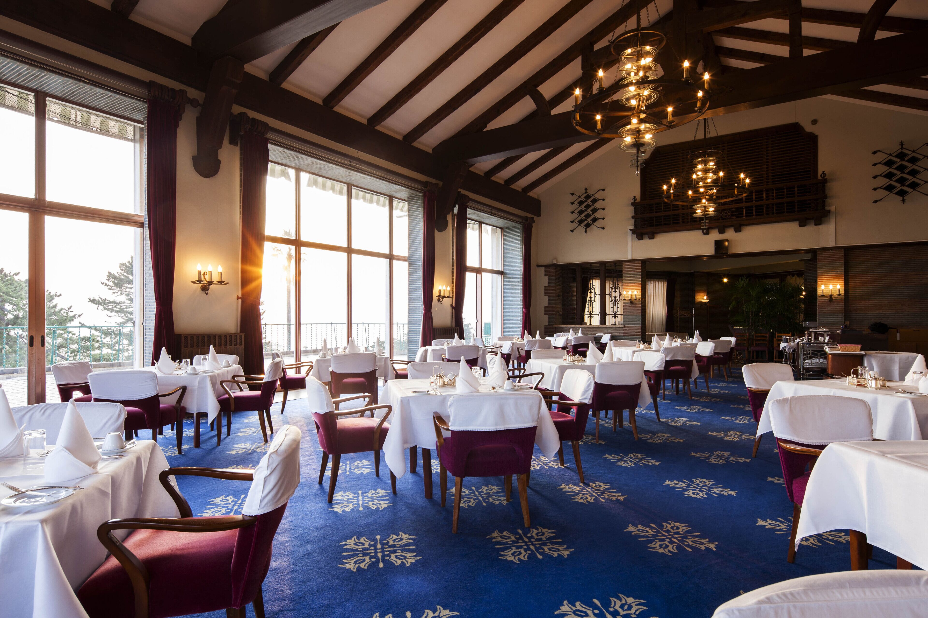 A dining area with tables set for service, large windows, and a chandelier.