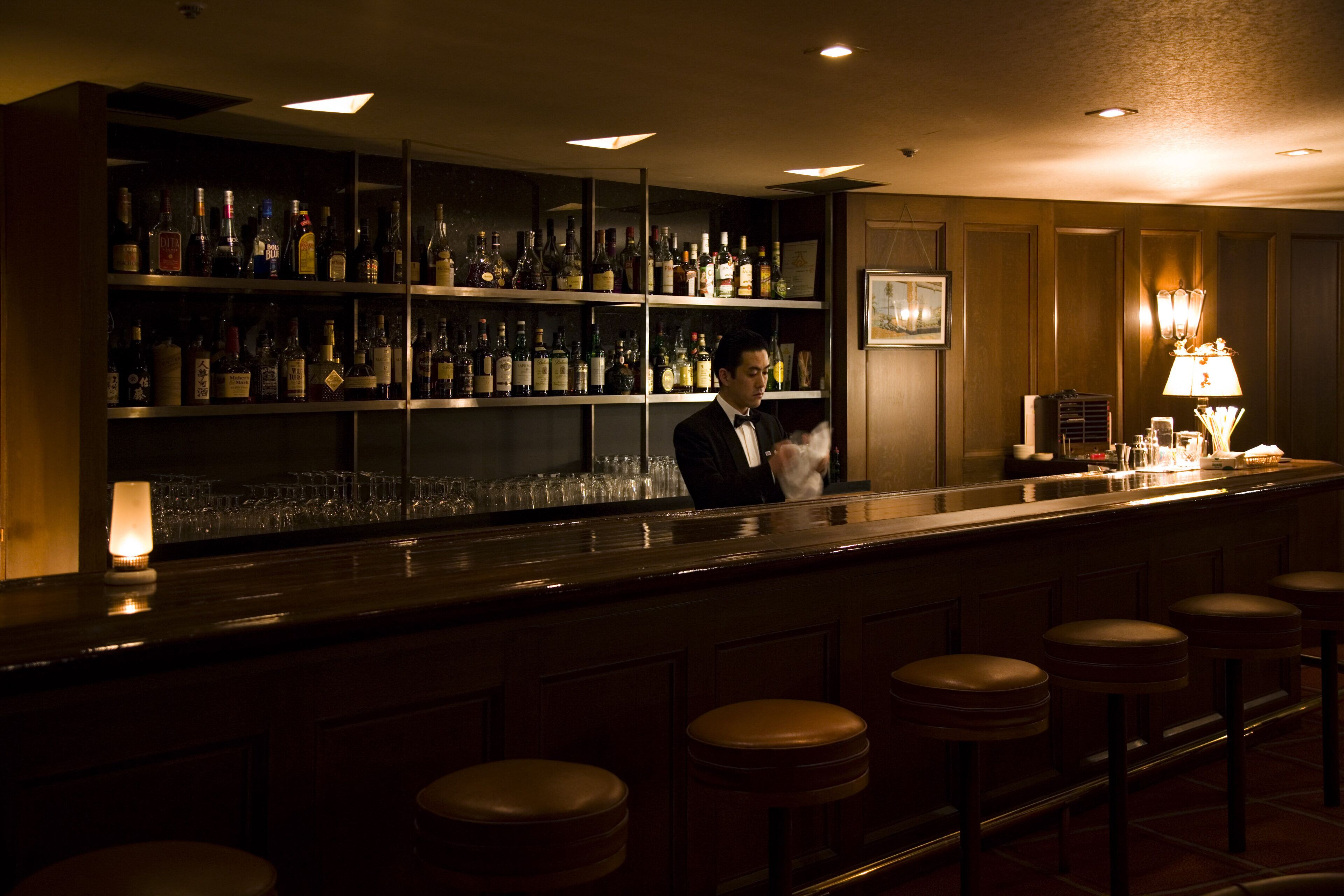 A bar area with a bartender, shelves of liquor bottles, and bar stools.
