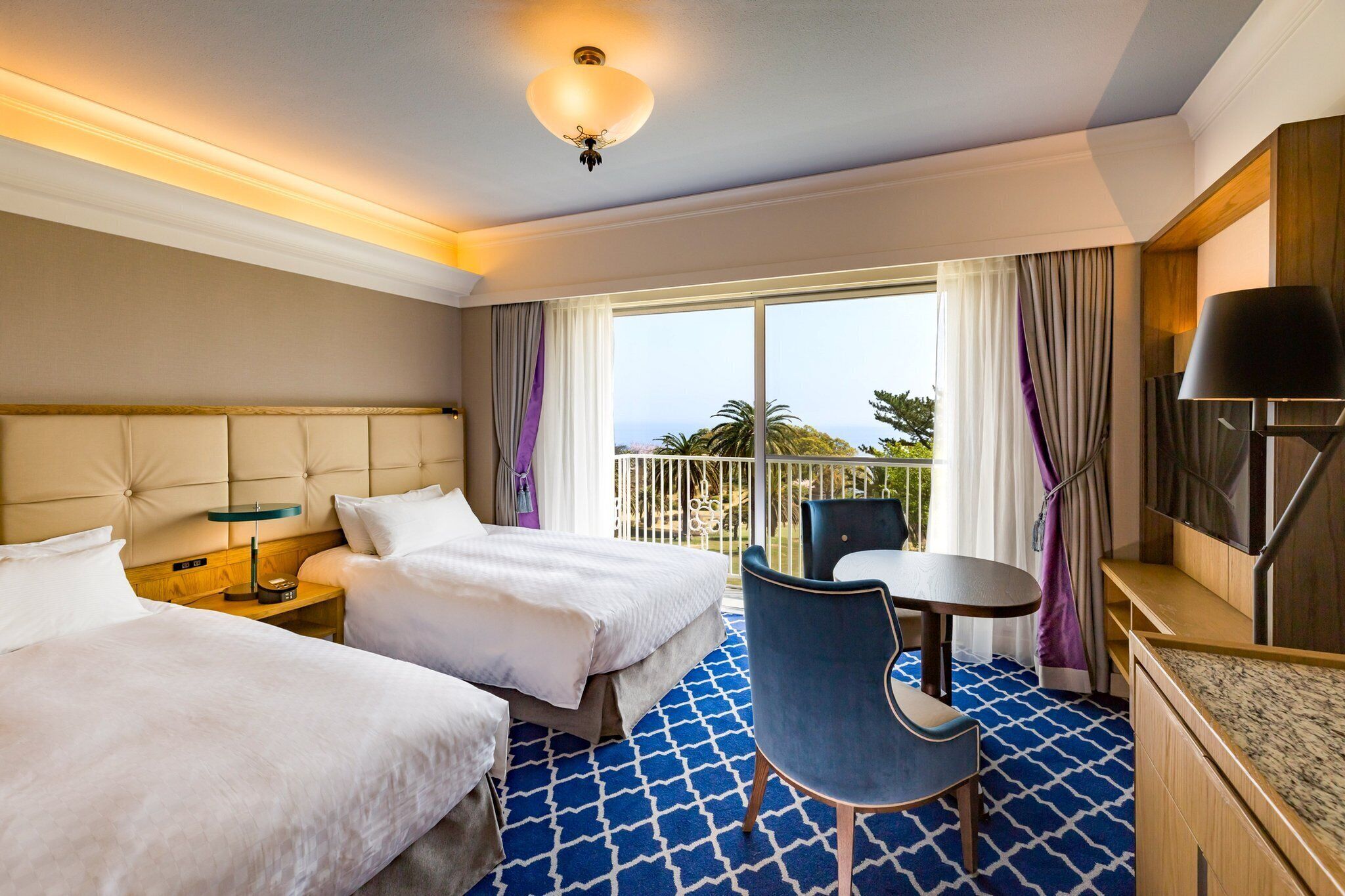 A hotel room with two beds, a balcony with a view, a small table with a chair, and a TV.