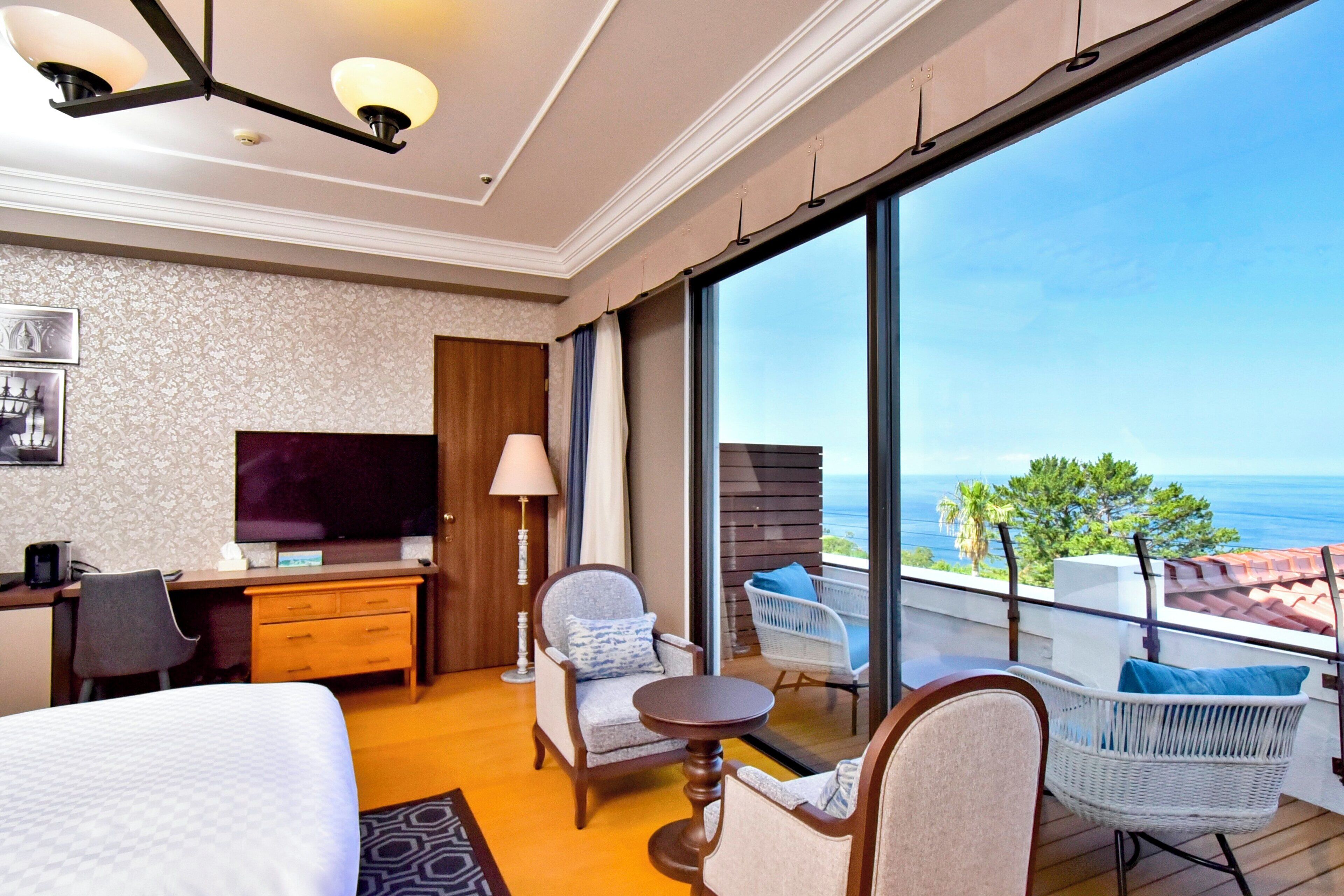 Habitación de hotel con cama, escritorio, televisor y balcón con sillas y vista al mar.