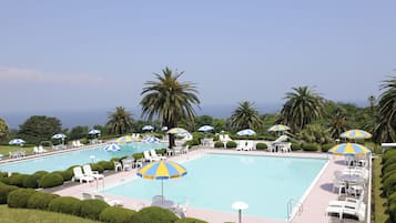 A large outdoor swimming pool with multiple lounge chairs and umbrellas, surrounded by neatly trimmed bushes and palm trees.