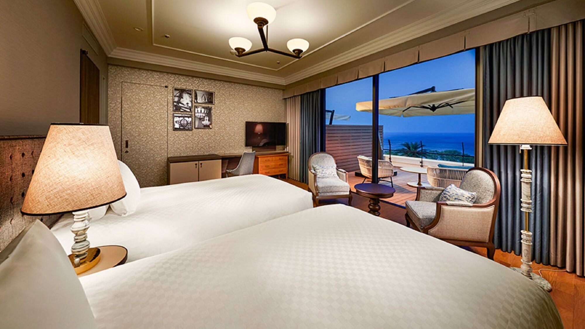 A hotel room with two beds, a desk, a TV, and a view of the ocean.