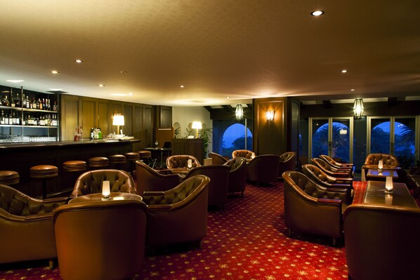 A bar area with leather chairs, a bar counter with bottles, and a view of the night sky through a window.