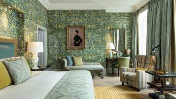 A hotel room with a large bed, a sofa, a chair, a desk with a lamp, and a painting on the wall.