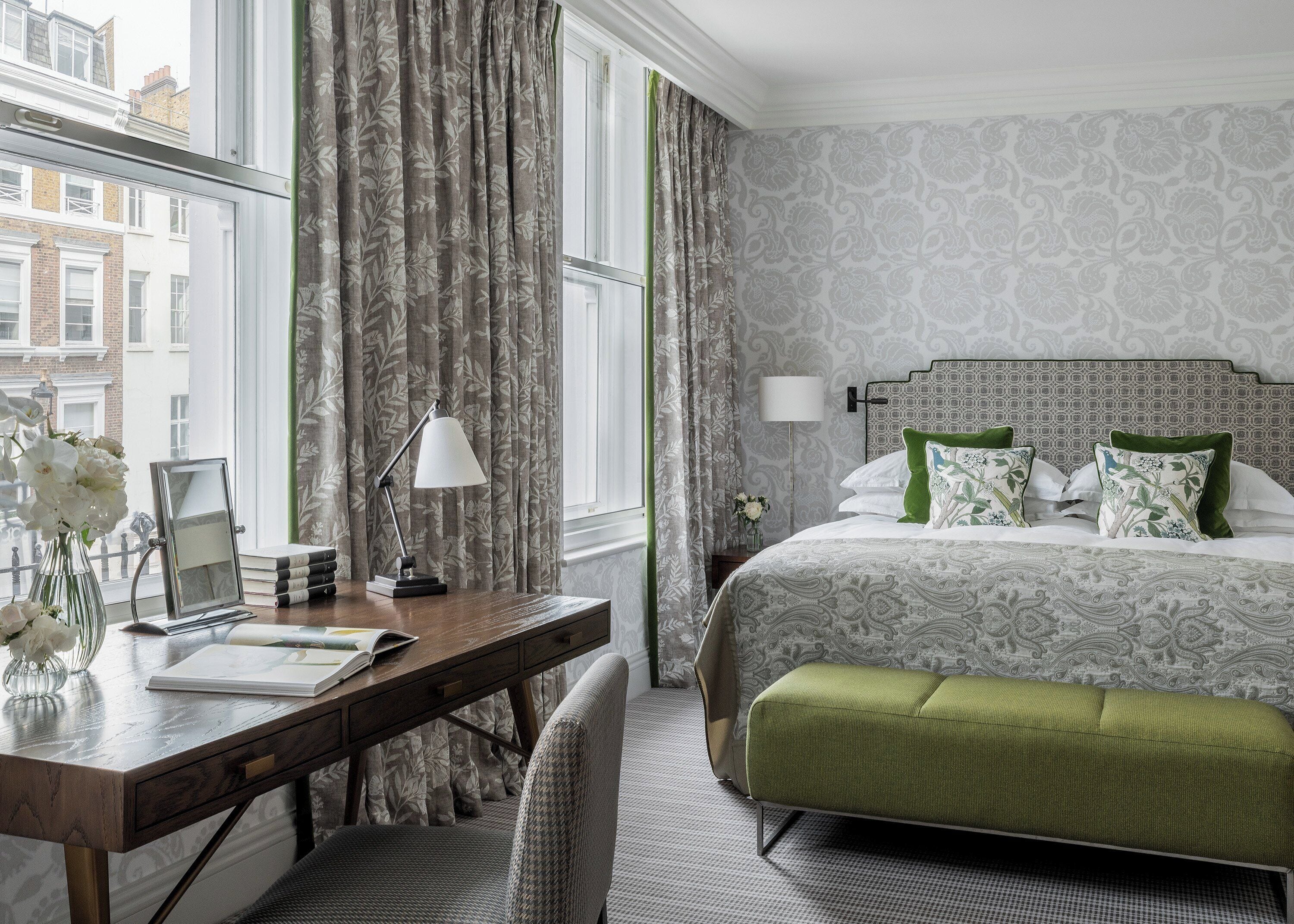 A hotel room with a bed, a desk with a lamp, a chair, and a view of buildings outside the window.