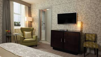 A hotel room with a bed, a green armchair, a small table with a lamp, a dark wooden cabinet, and a TV mounted on the wall.