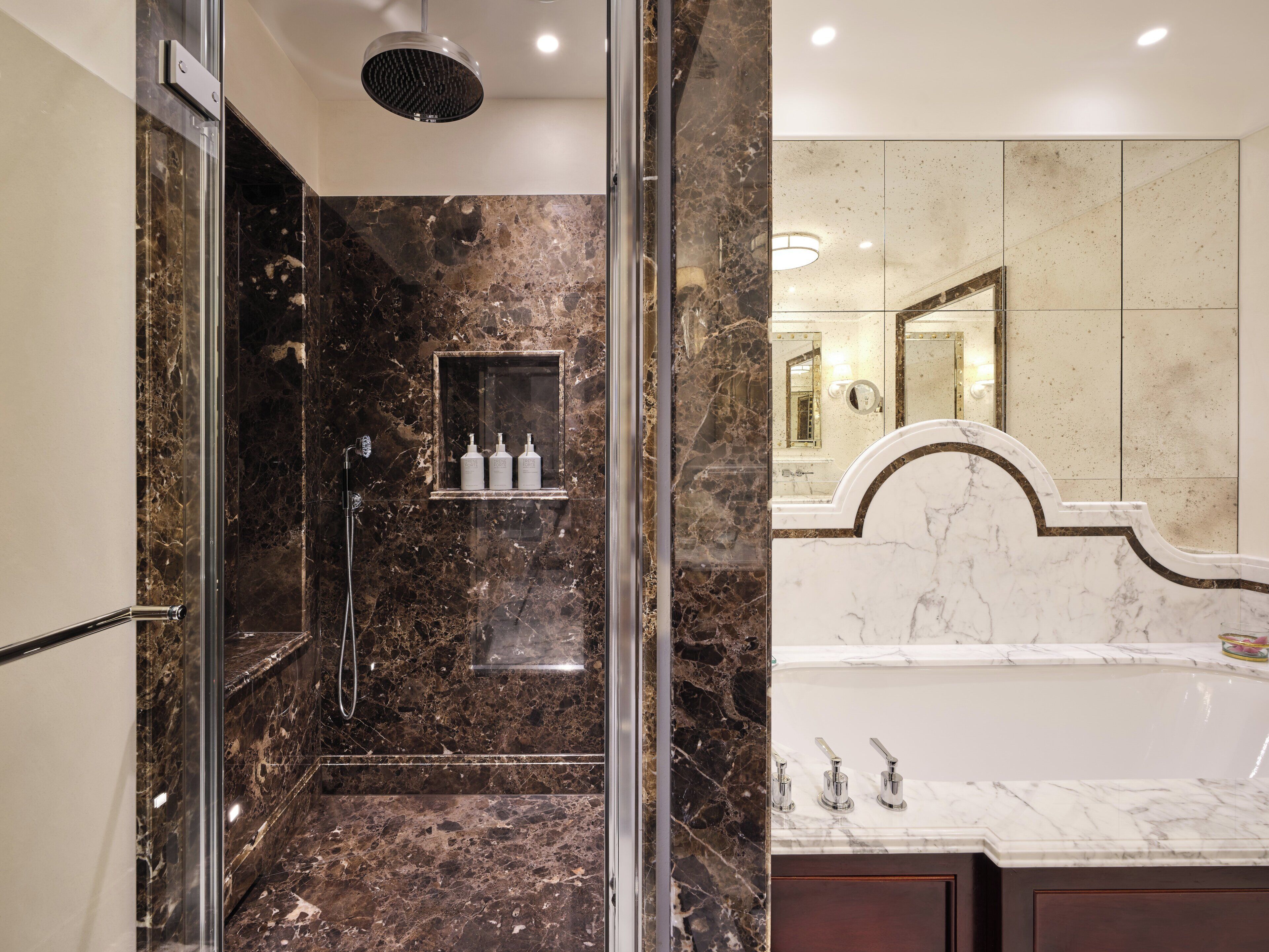 A modern bathroom with a glass-enclosed shower, marble walls, a built-in shelf with toiletries, and a separate bathtub area.