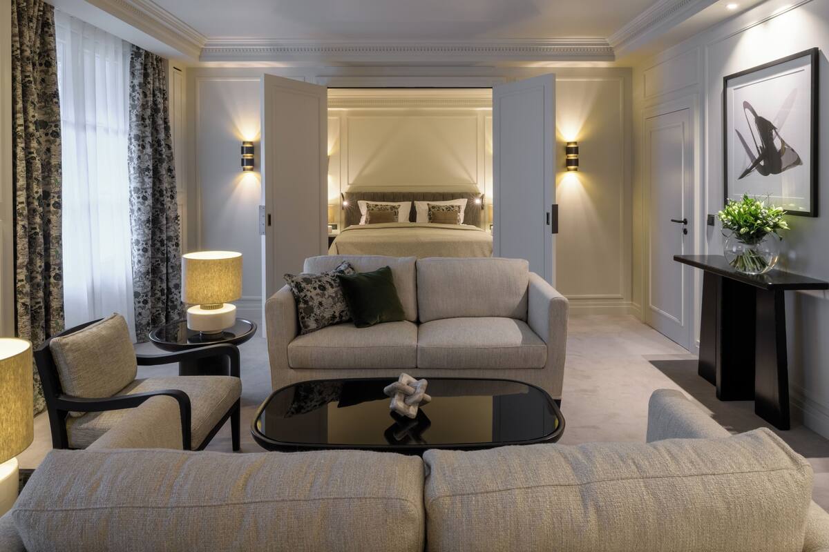 A hotel room with a sofa, armchair, and a coffee table. There is a bed with a canopy and a wall-mounted lamp.