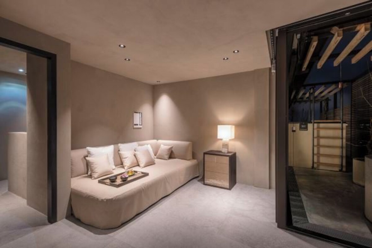 A modern hotel room with a sofa, a bedside table with a lamp, and a large mirror reflecting the room.