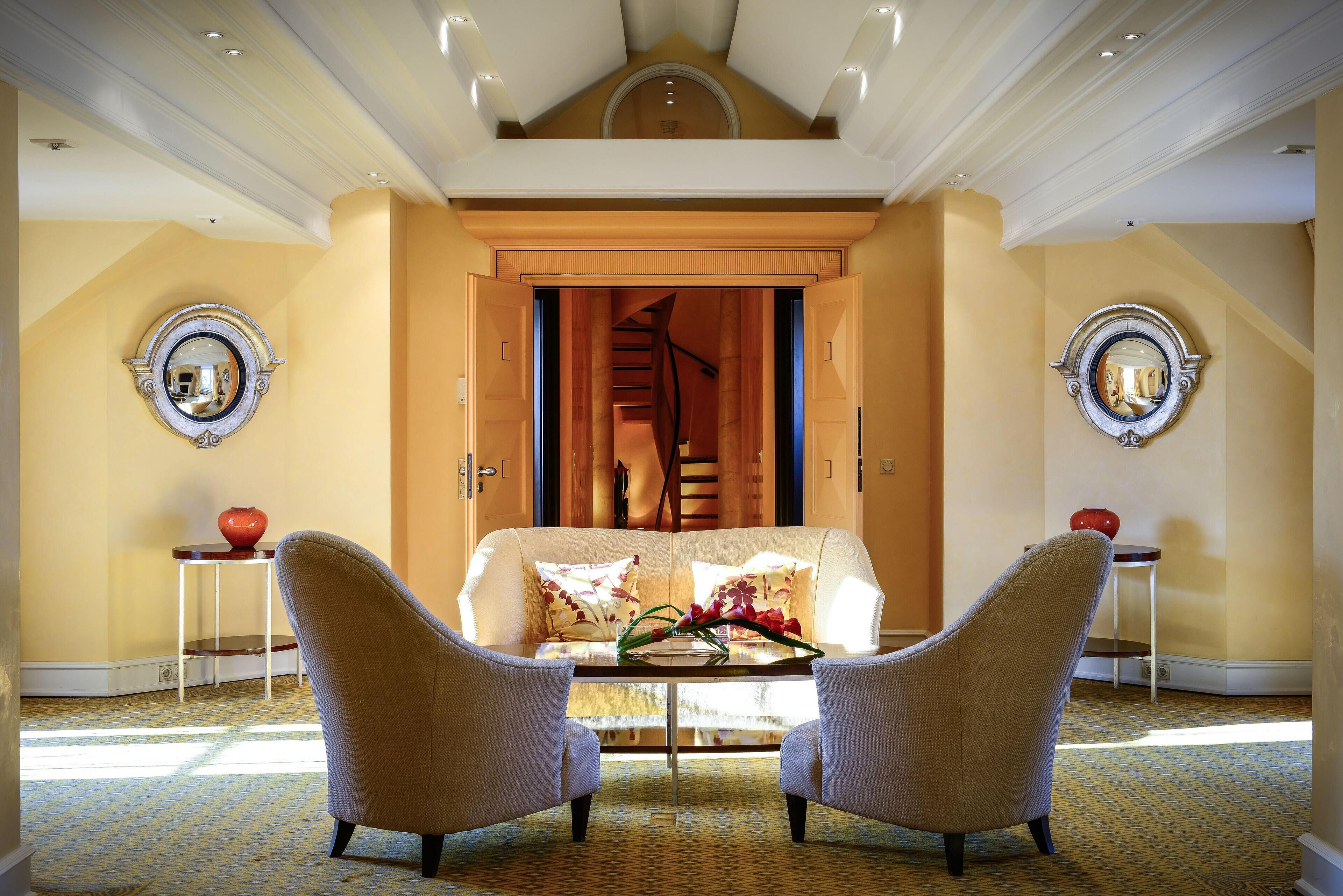 A spacious room with a high ceiling, a central seating area with two armchairs and a sofa, a small table with a vase, and a staircase leading to an upper level.