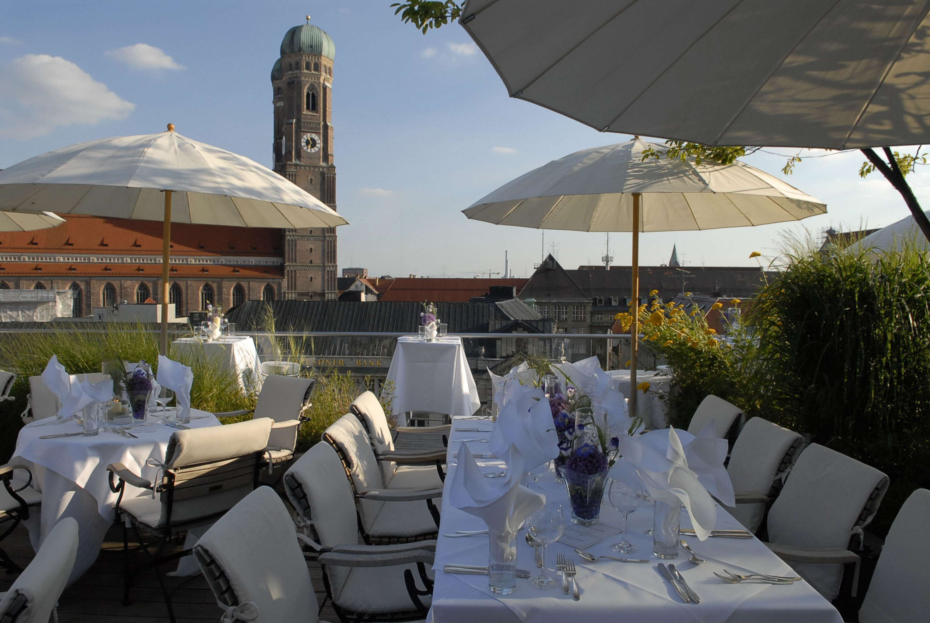 An outdoor dining area with tables set for two, featuring white tablecloths and chairs, with umbrellas providing shade.