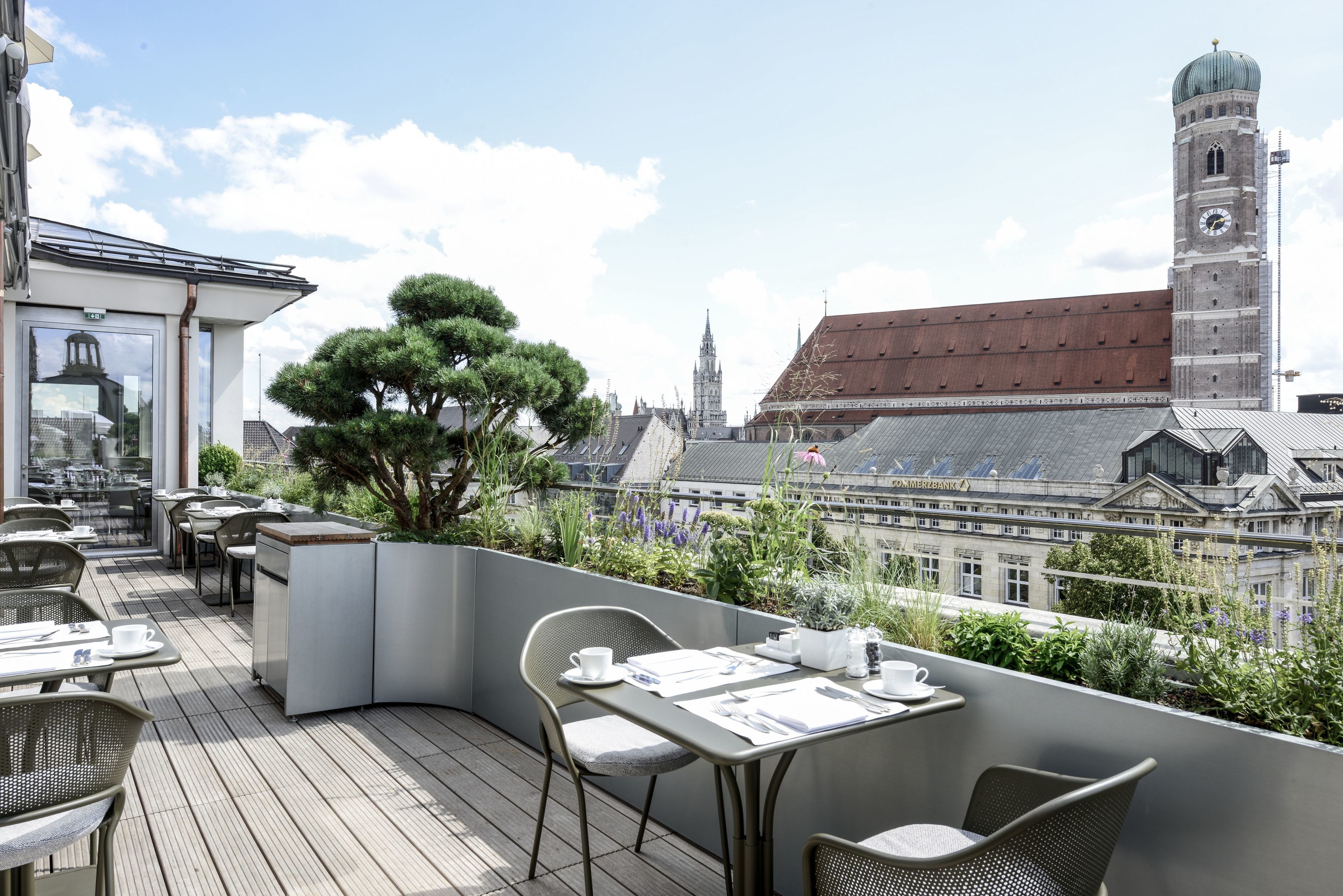 Terraço no telhado com mesas e cadeiras, uma grande árvore e vista para edifícios históricos e um campanário.