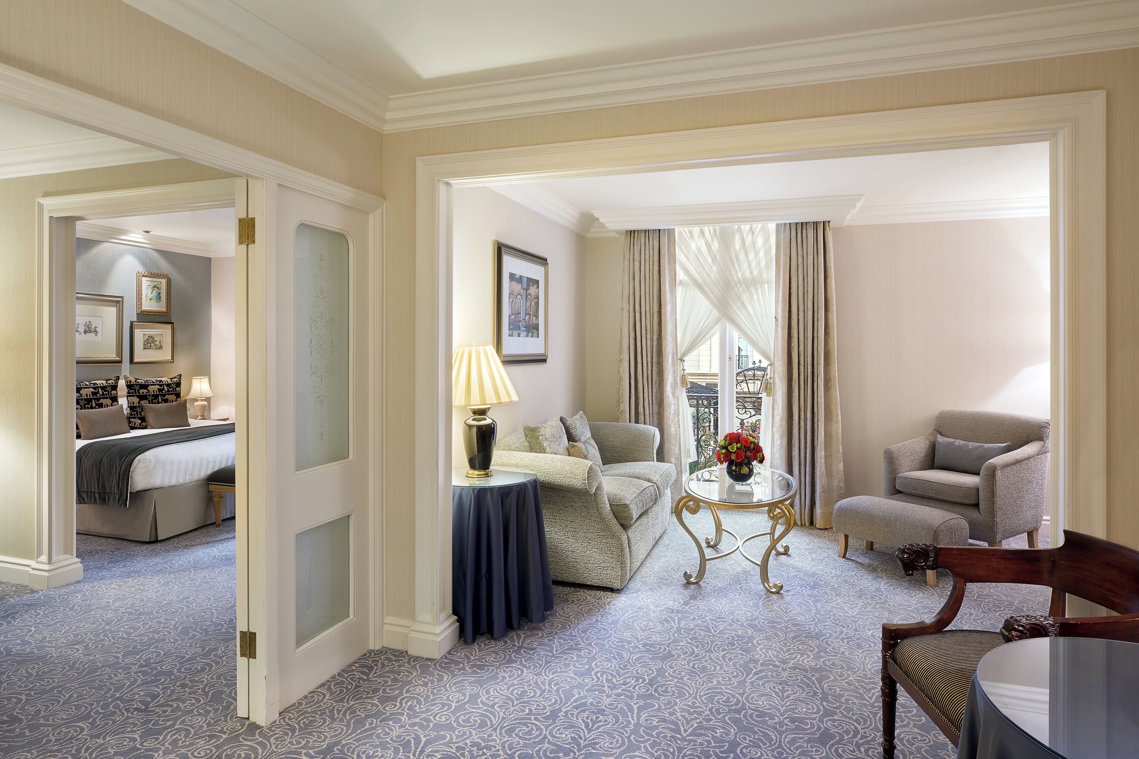 A hotel room with a bed, a sitting area with a sofa and armchairs, a small table with a lamp, and a balcony with a railing.
