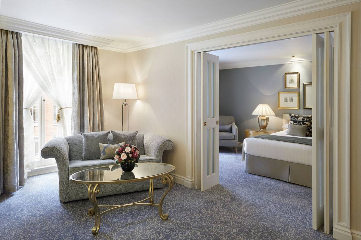 Et hotelværelse med en sofa, et glasbord med en blomstervase, en seng med et mønstret sengetæppe og et skrivebord med en lampe.