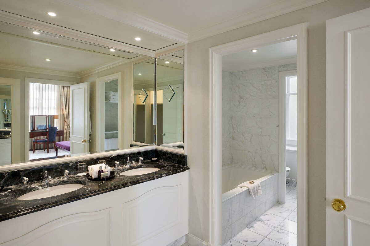 Un baño con lavamanos doble, un espejo grande, una bañera y una puerta que da a otra habitación.