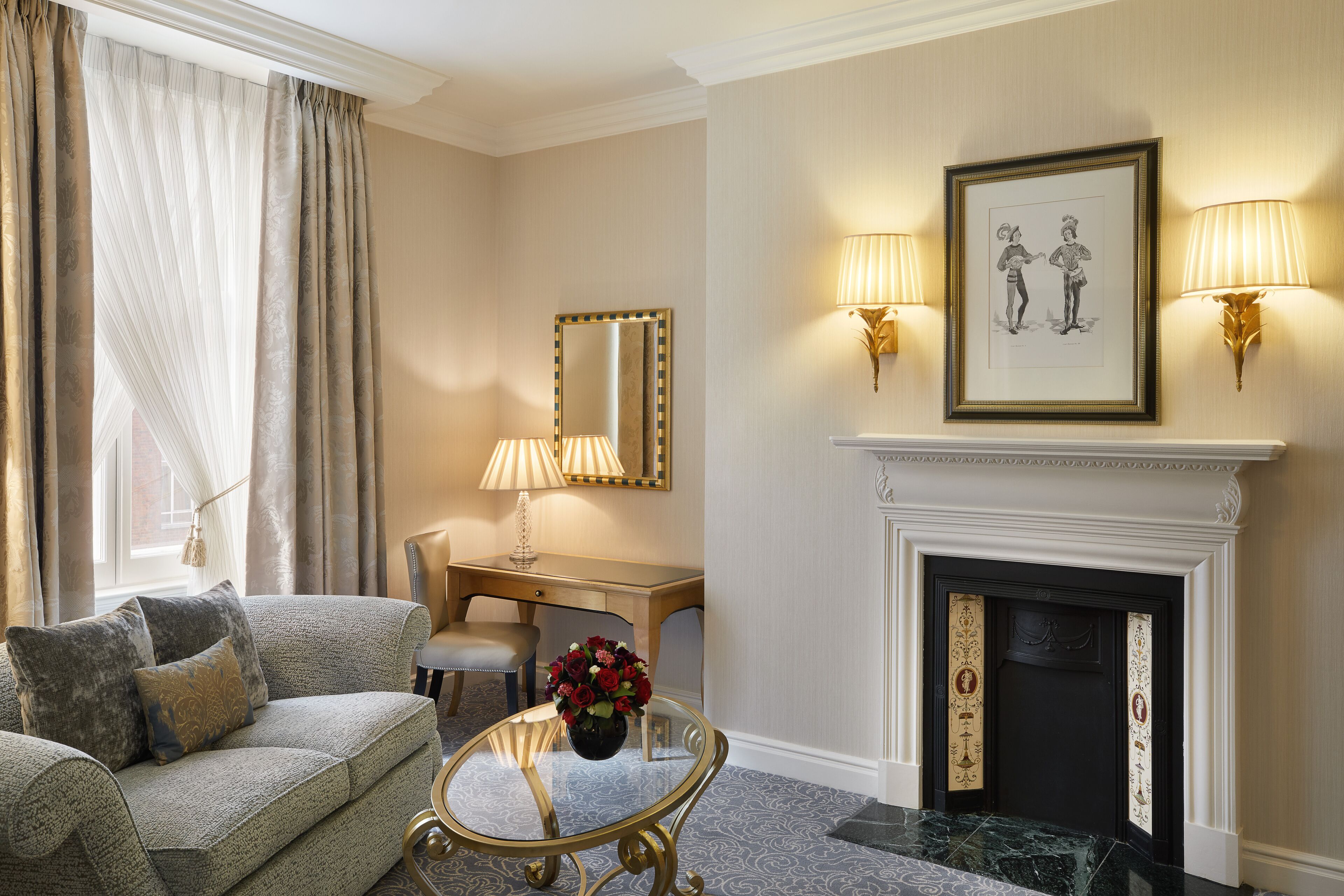 A living room with a sofa, a table with a vase of flowers, a desk, a chair, a fireplace, and wall-mounted lamps.