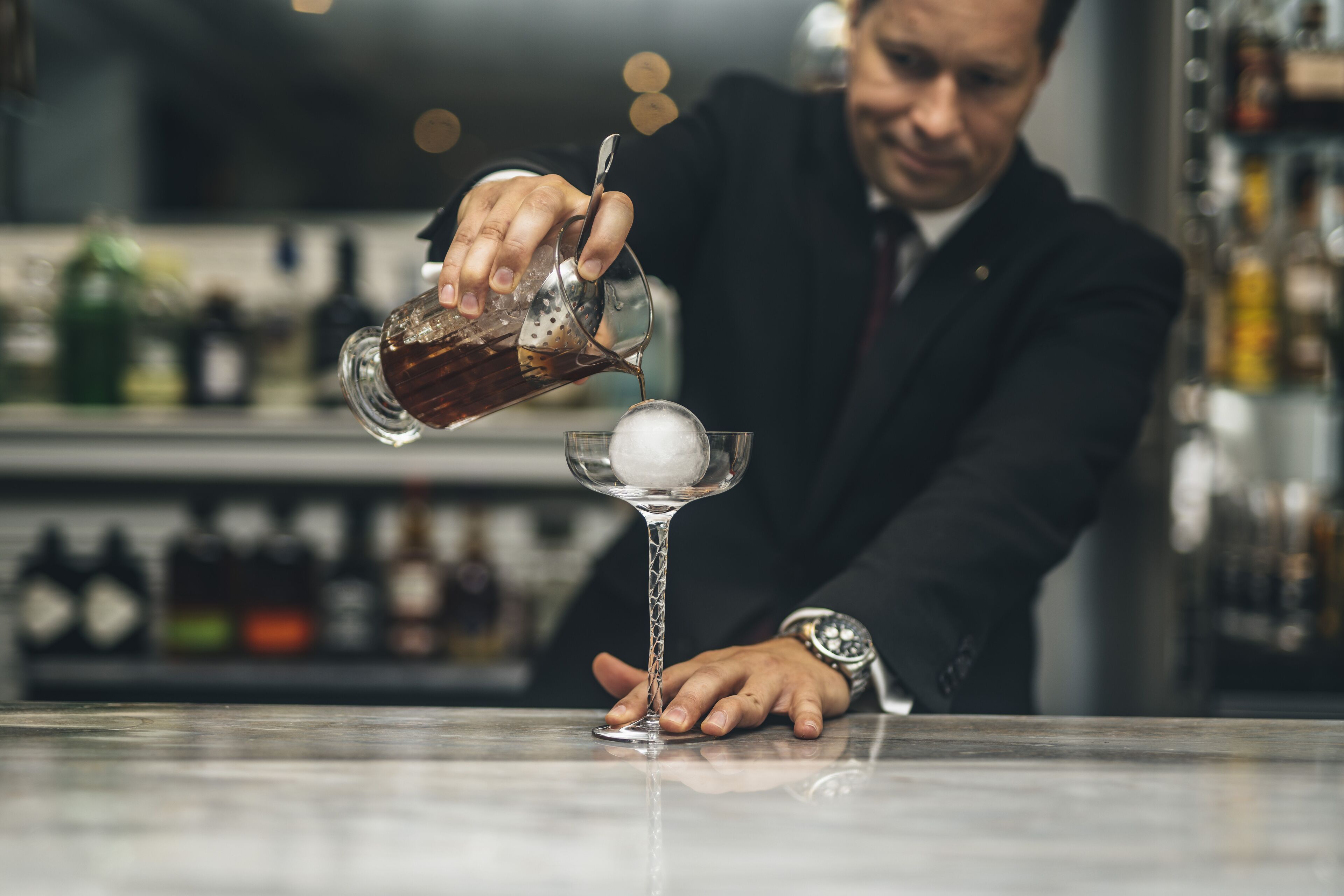 Ein Barkeeper gießt ein Getränk in ein Glas mit einem großen Eiswürfel.
