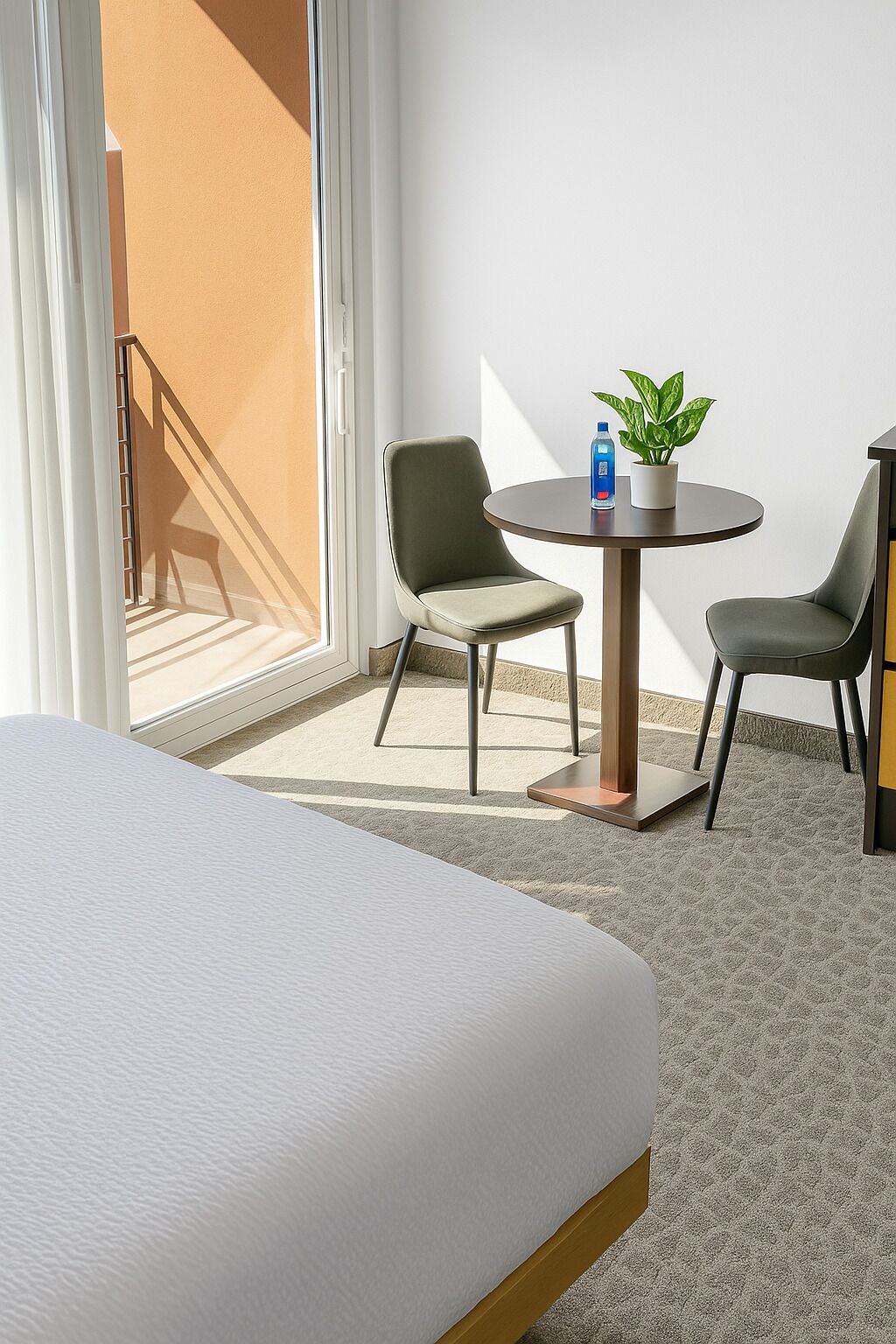 Quarto de hotel com cama, duas cadeiras, uma mesinha com uma planta e uma garrafa, e vista para o exterior.