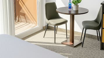 Quarto de hotel com cama, duas cadeiras, uma mesinha com uma planta e uma garrafa, e vista para o exterior.