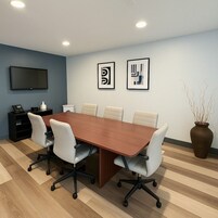 A conference room with a long wooden table, six chairs, a TV on the wall, and framed abstract art.