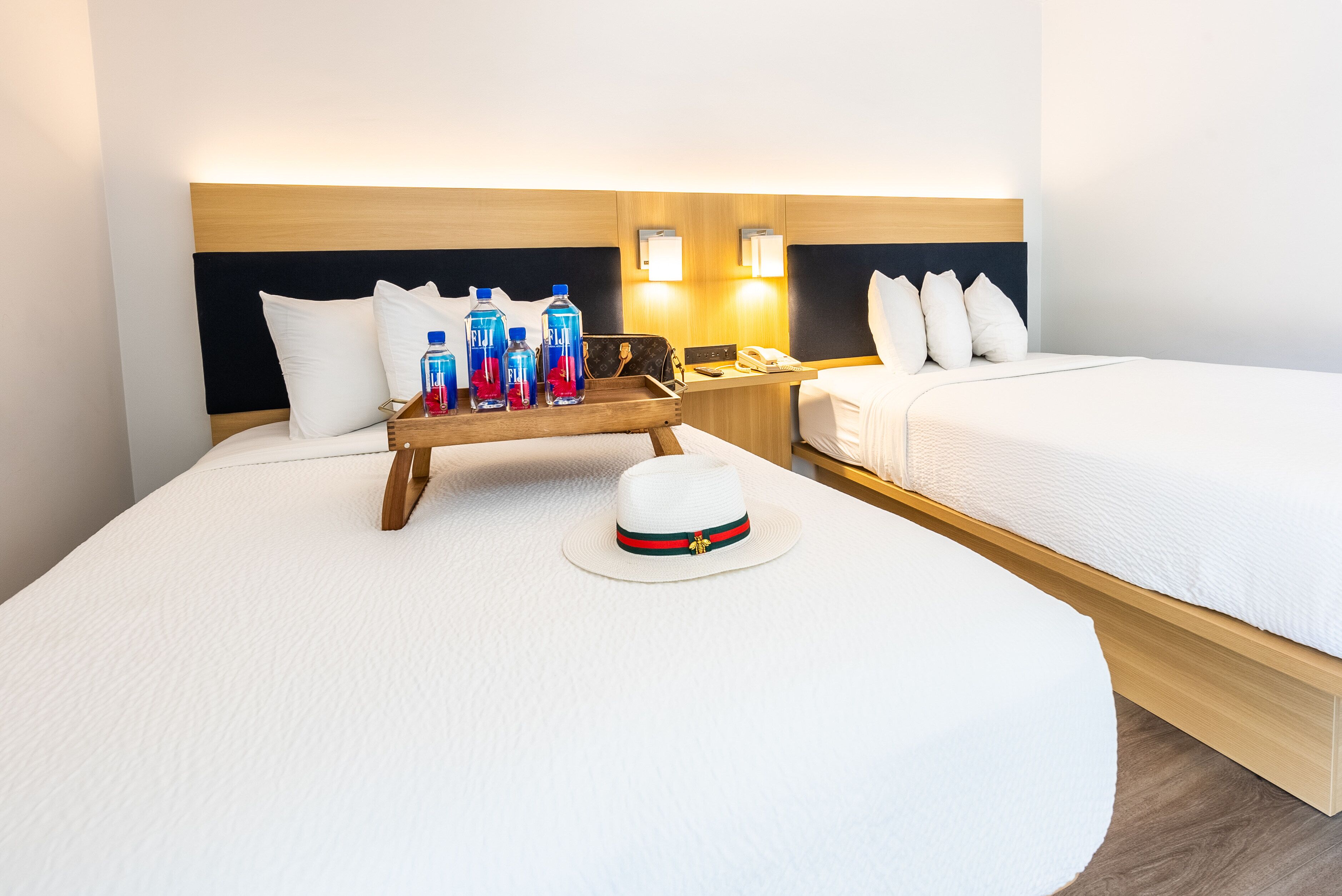 A hotel room with two beds, a wooden headboard, bedside tables, and a tray with water bottles.
