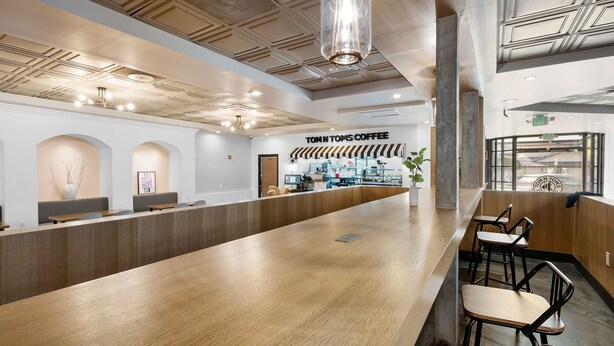 A modern coffee shop interior with a long wooden counter, black bar stools, and a ceiling with recessed lighting. - The Live Hotel (Los Angeles)