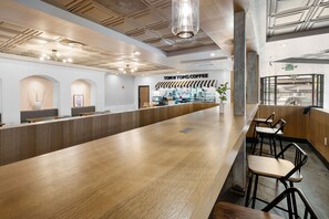 A modern coffee shop interior with a long wooden counter, black bar stools, and a ceiling with recessed lighting. - The Live Hotel (Los Angeles)