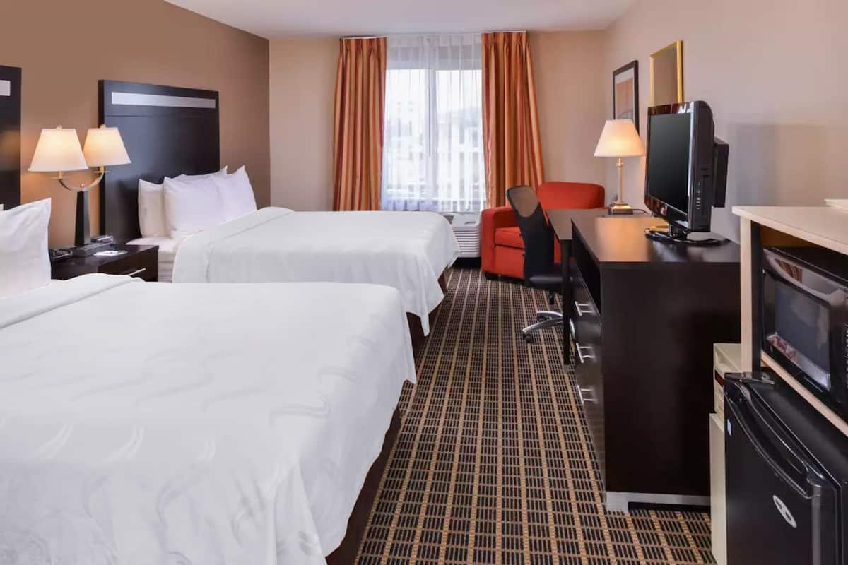Una habitación de hotel con dos camas, un escritorio con computadora, un televisor y una silla roja.