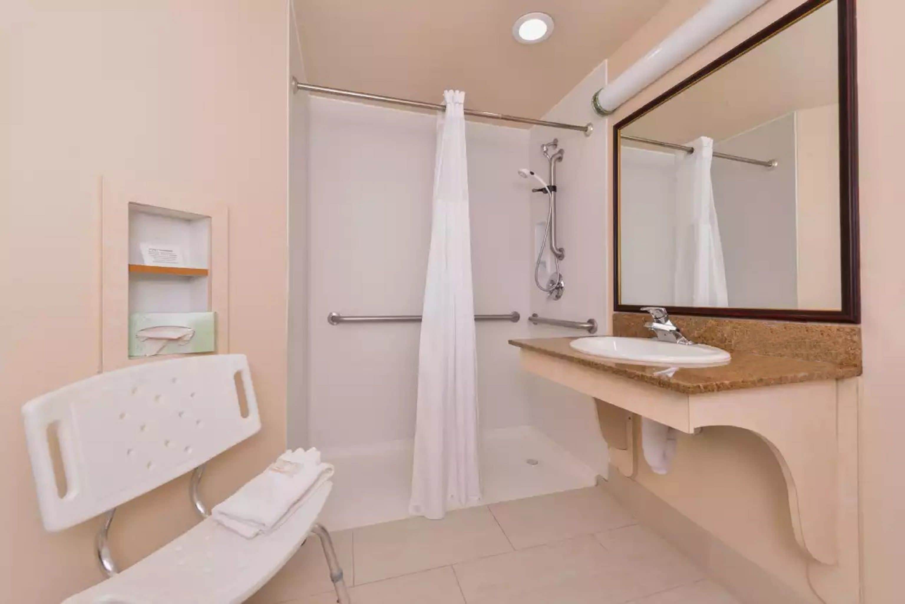 Un baño con ducha, lavamanos, espejo y banco.