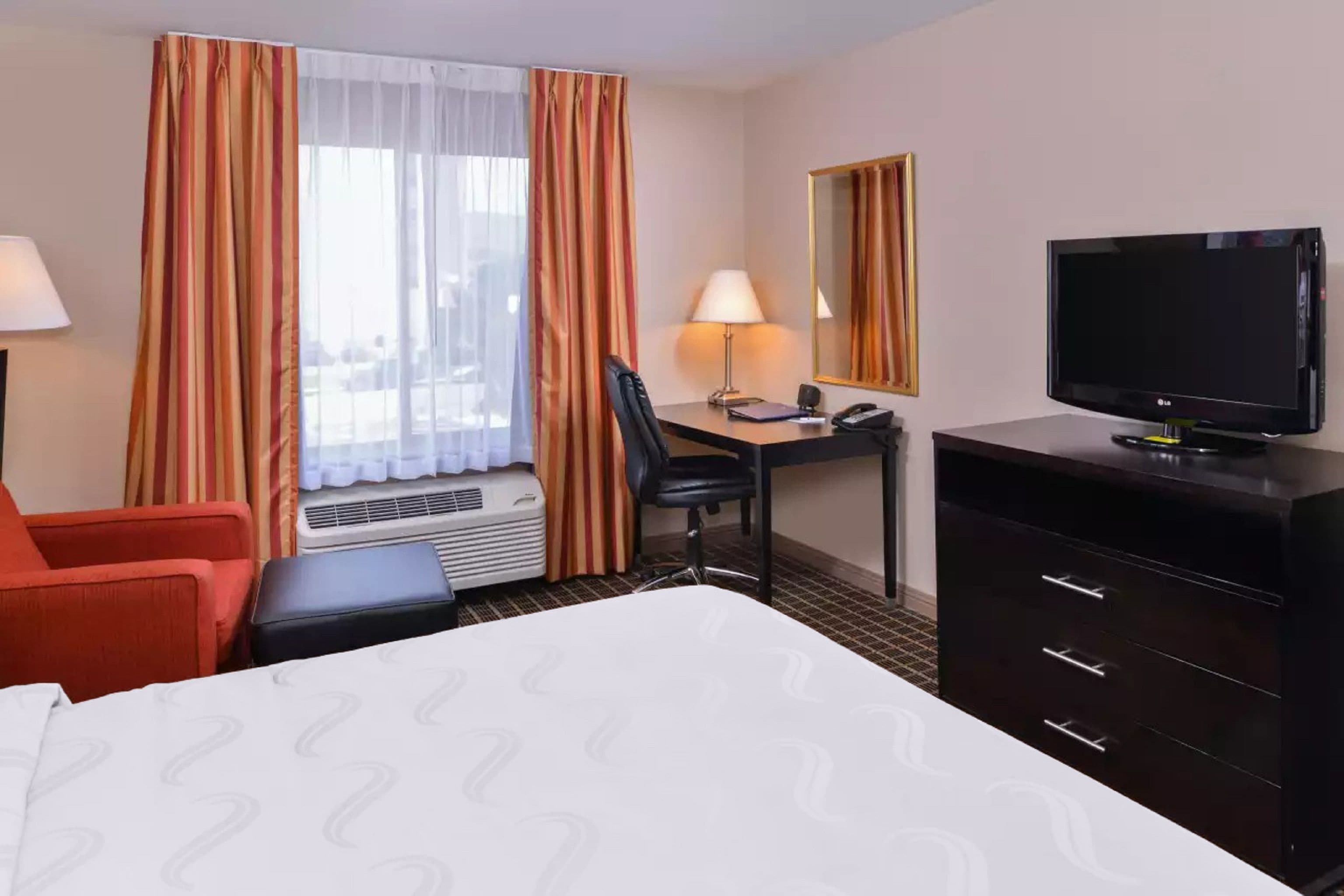 A hotel room with a bed, a desk with a chair, a television, a dresser, and a window with curtains.