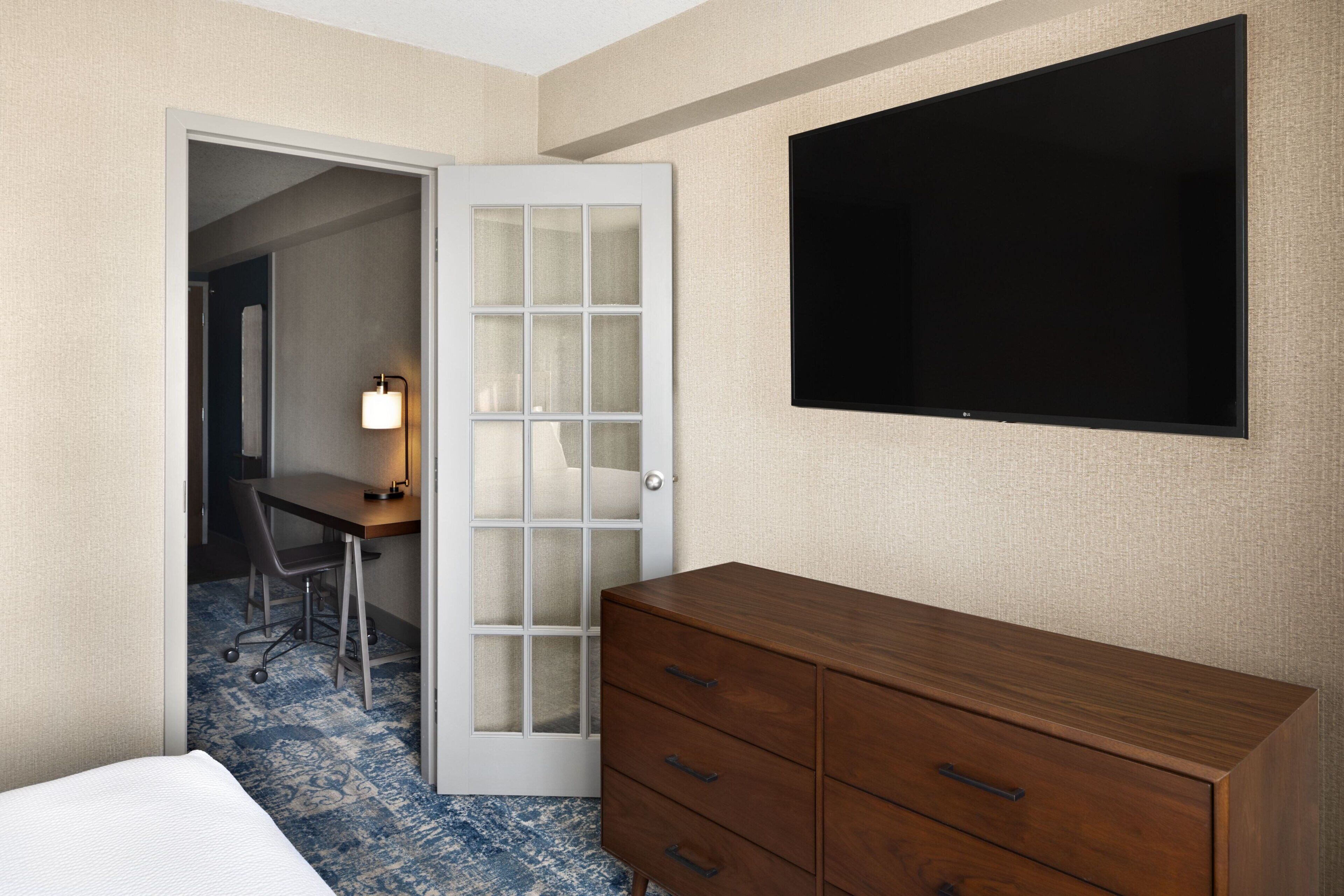 Habitación de hotel con una cama, un aparador de madera, un escritorio con una silla, una lámpara y un televisor montado en la pared.