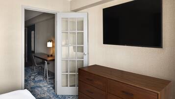 Habitación de hotel con una cama, un aparador de madera, un escritorio con una silla, una lámpara y un televisor montado en la pared.