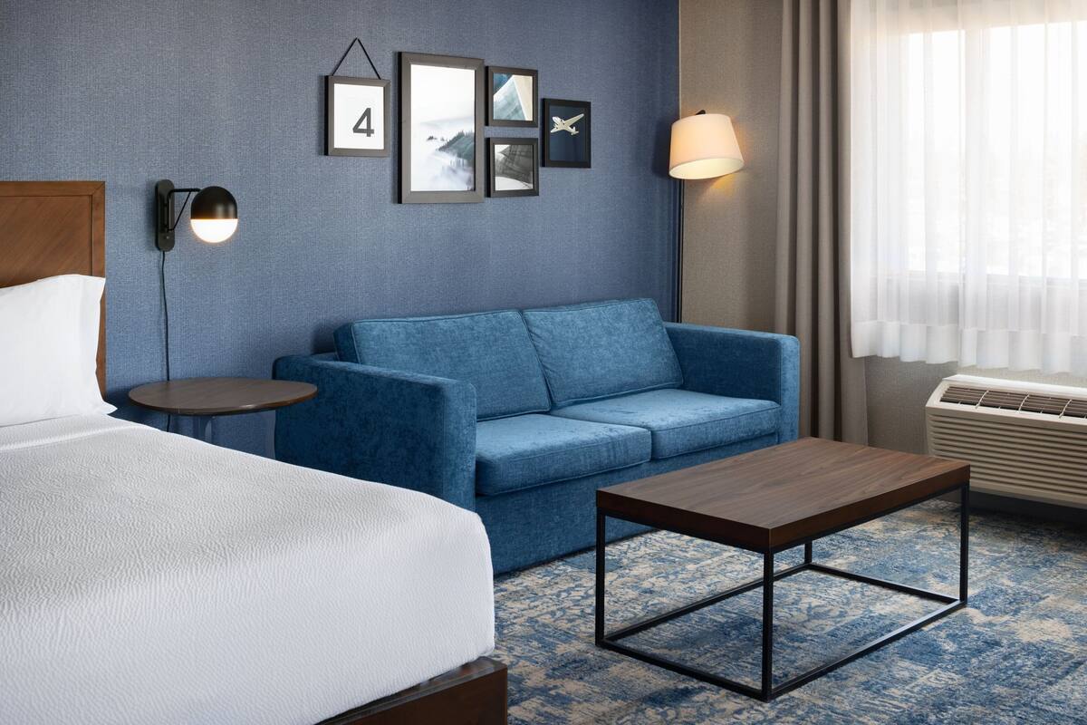 A hotel room with a bed, a blue sofa, a small table, and a wall-mounted lamp.