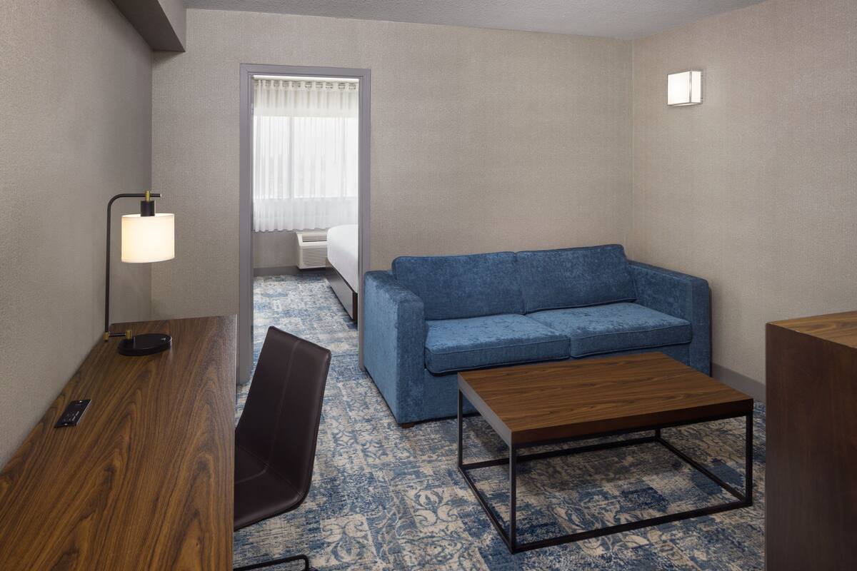 A hotel room with a wooden desk, a blue sofa, a wooden coffee table, a black chair, and a window with curtains.