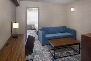 Habitación de hotel con un escritorio de madera, un sofá azul, una mesa de centro de madera, una silla negra y una ventana con cortinas.
