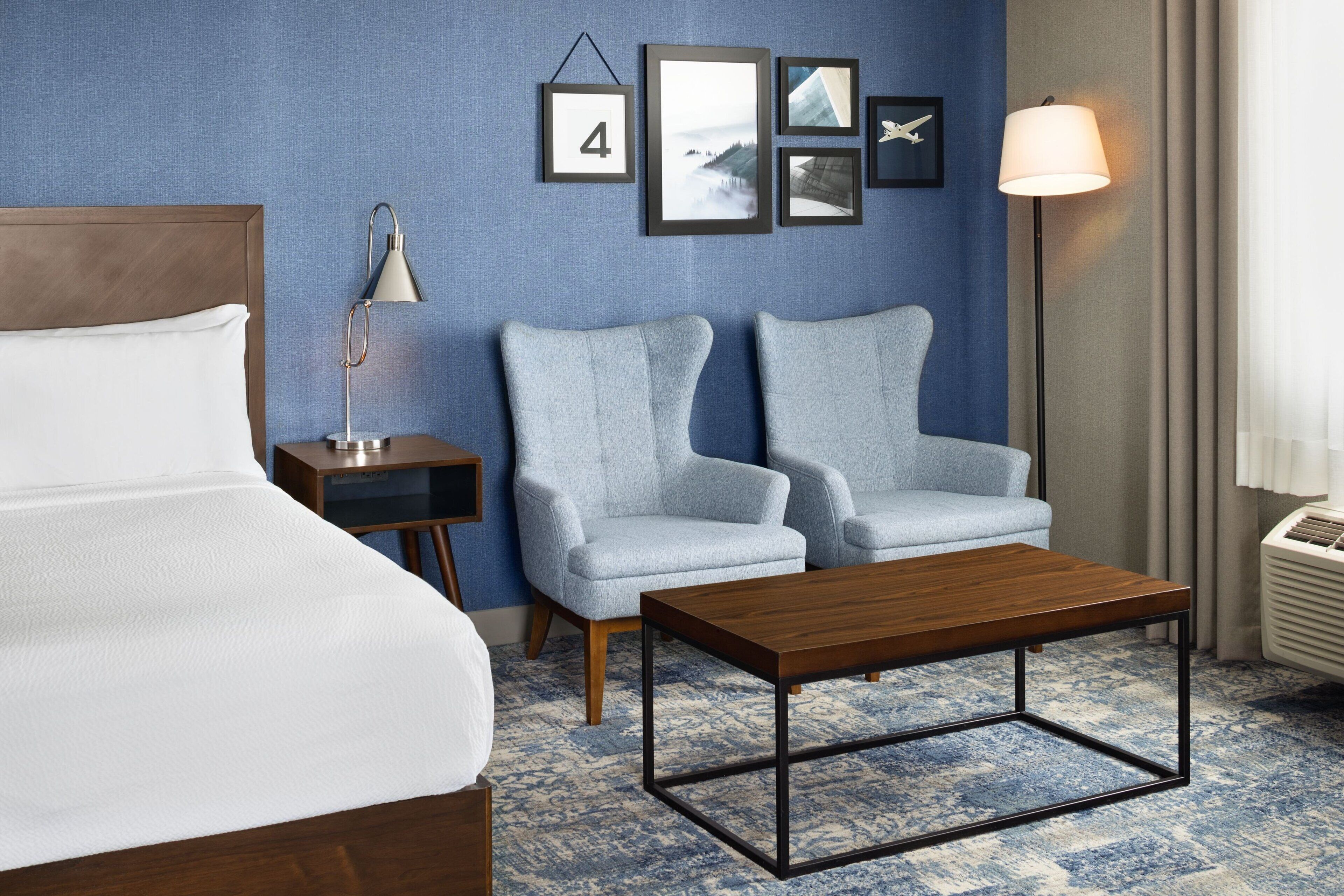 A hotel room with a bed, two armchairs, a wooden coffee table, a nightstand, a lamp, and framed pictures on the wall.