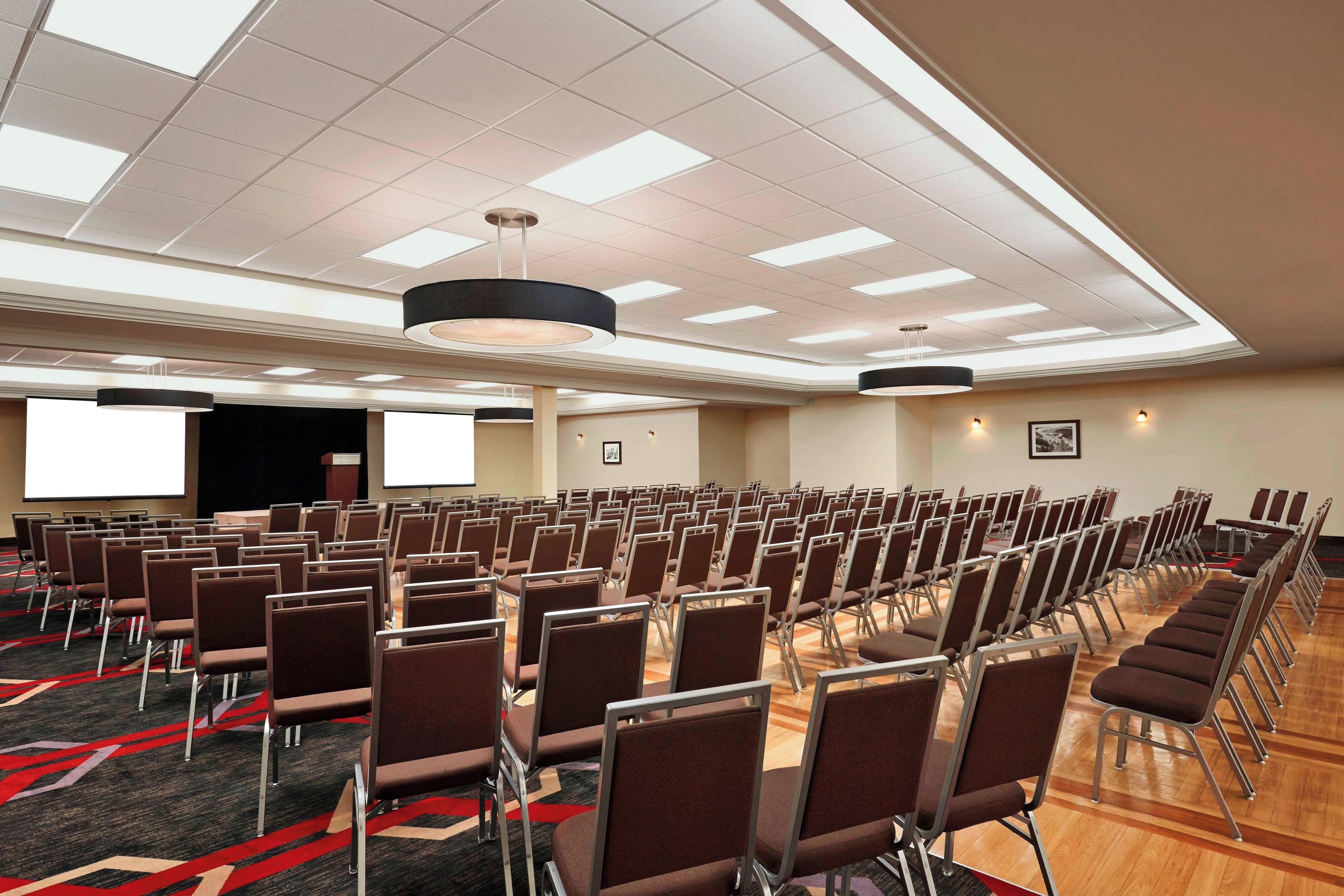 Sala de conferências com fileiras de cadeiras, uma tela de projetor e um palco.
