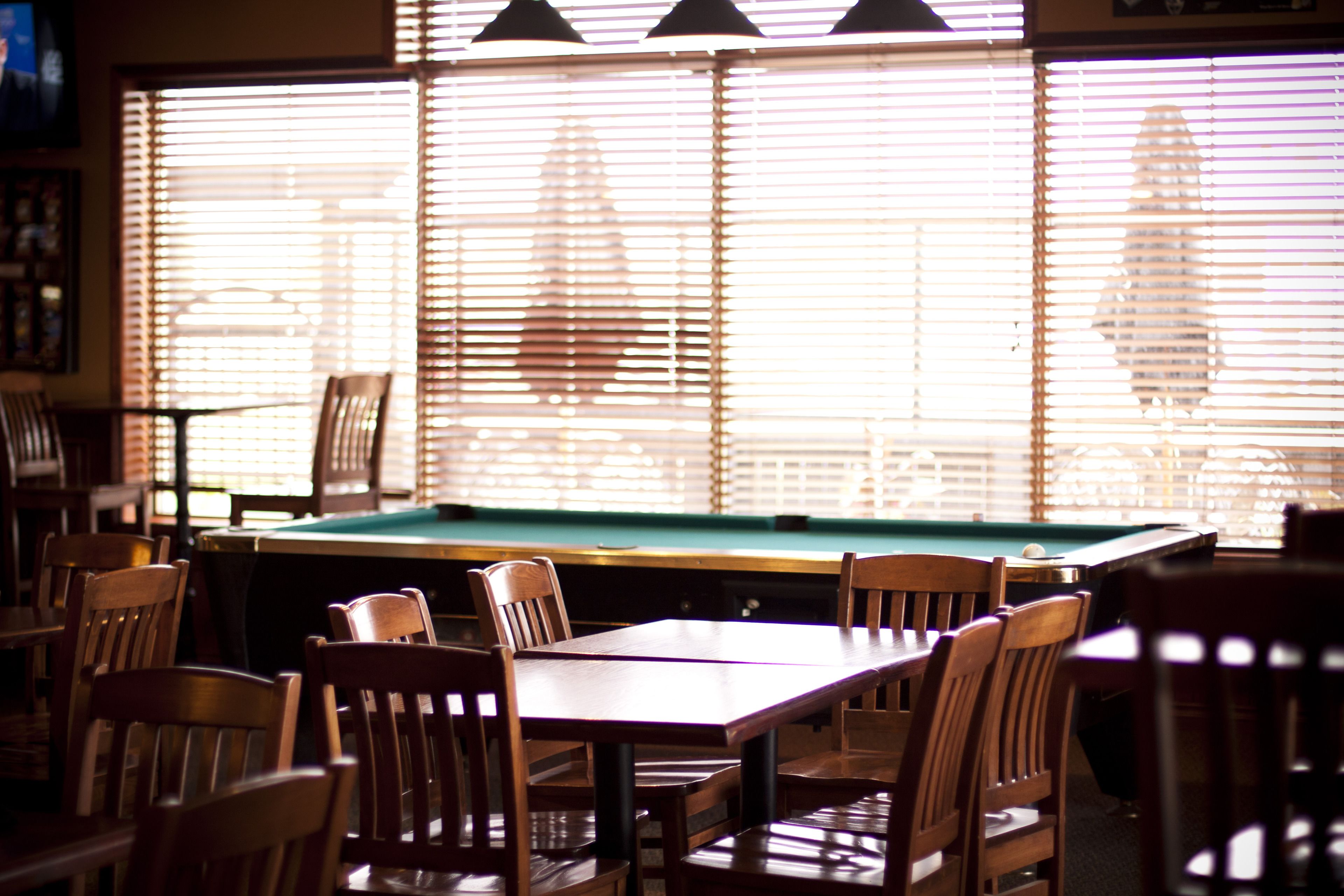 A pool table with chairs around it, a television mounted on the wall, and a window with blinds.