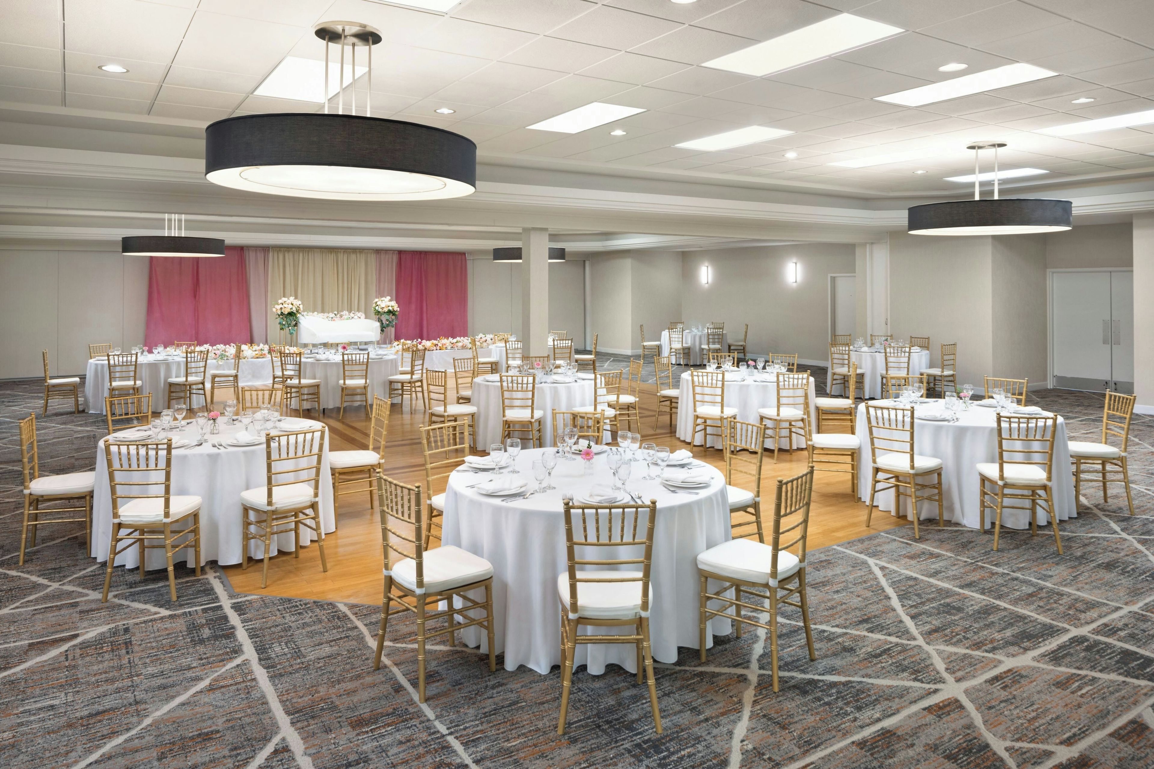 Une grande salle de conférence dotée de nombreuses tables rondes, chacune recouverte d’une nappe blanche et entourée de chaises dorées, le tout disposé sur un tapis à motifs.