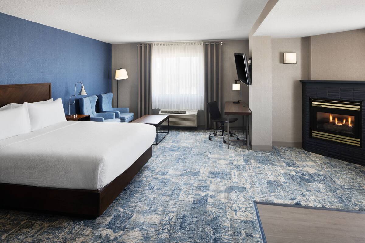 A hotel room with a large bed, a desk, a chair, a television, and a fireplace.