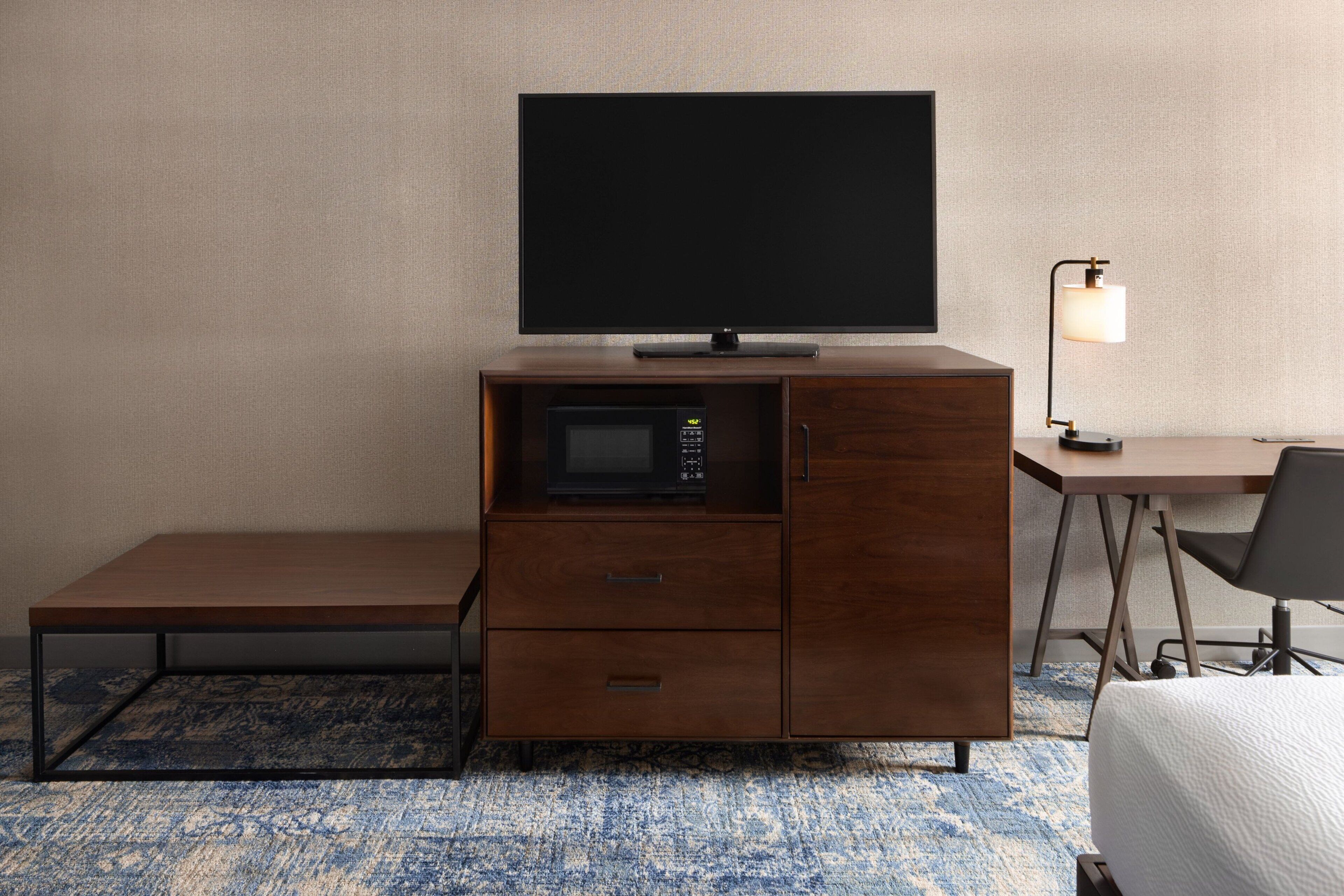 A compact hotel room with a flat-screen TV on a wooden cabinet, a microwave, a desk with a chair, and a bedside lamp.