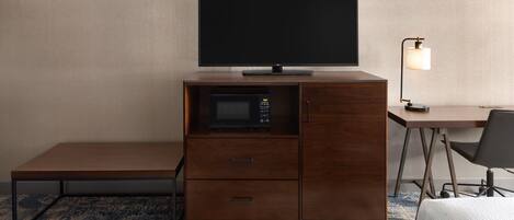 A compact hotel room with a flat-screen TV on a wooden cabinet, a microwave, a desk with a chair, and a bedside lamp.