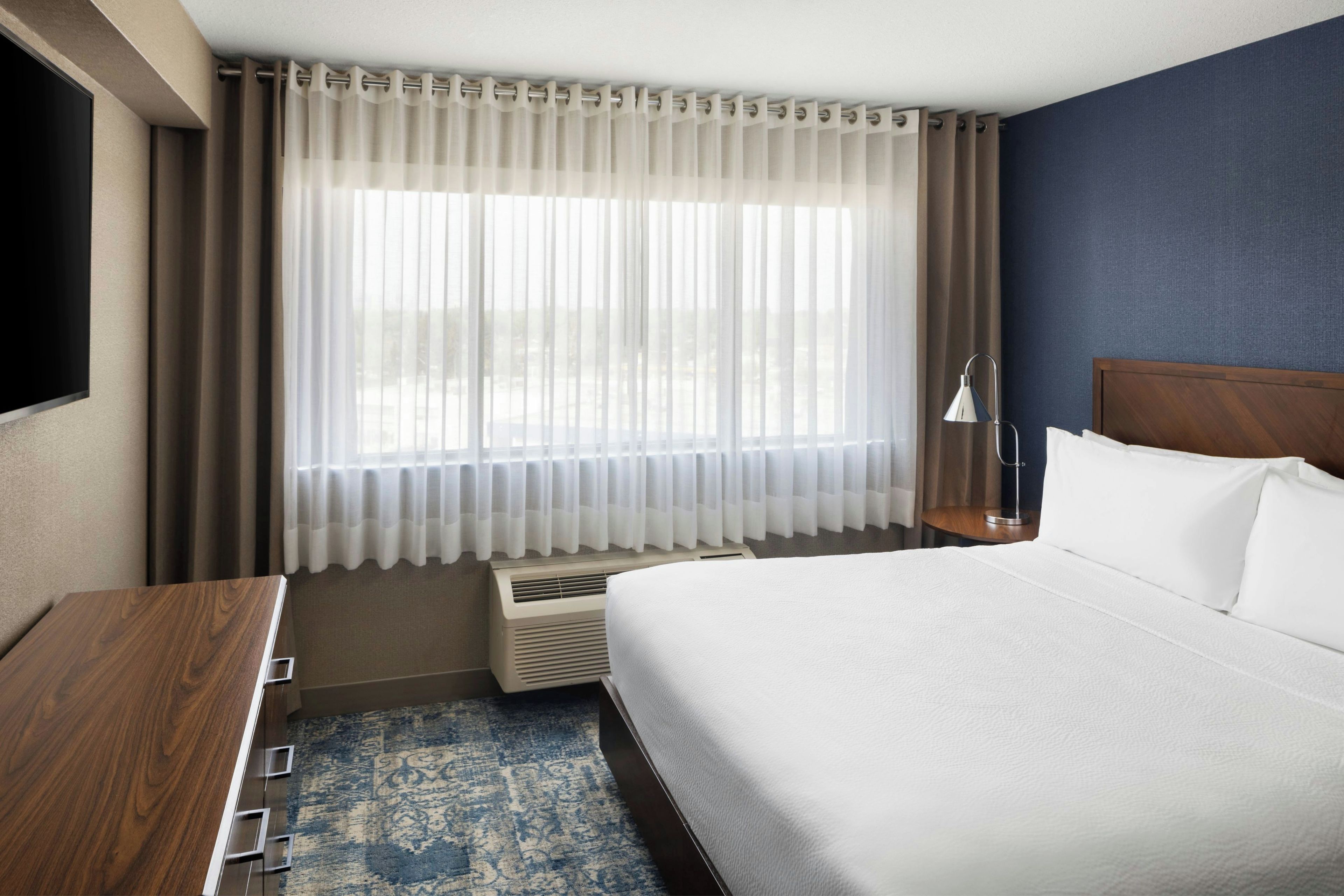 A hotel room with a bed, a desk, a lamp, and a window with curtains.