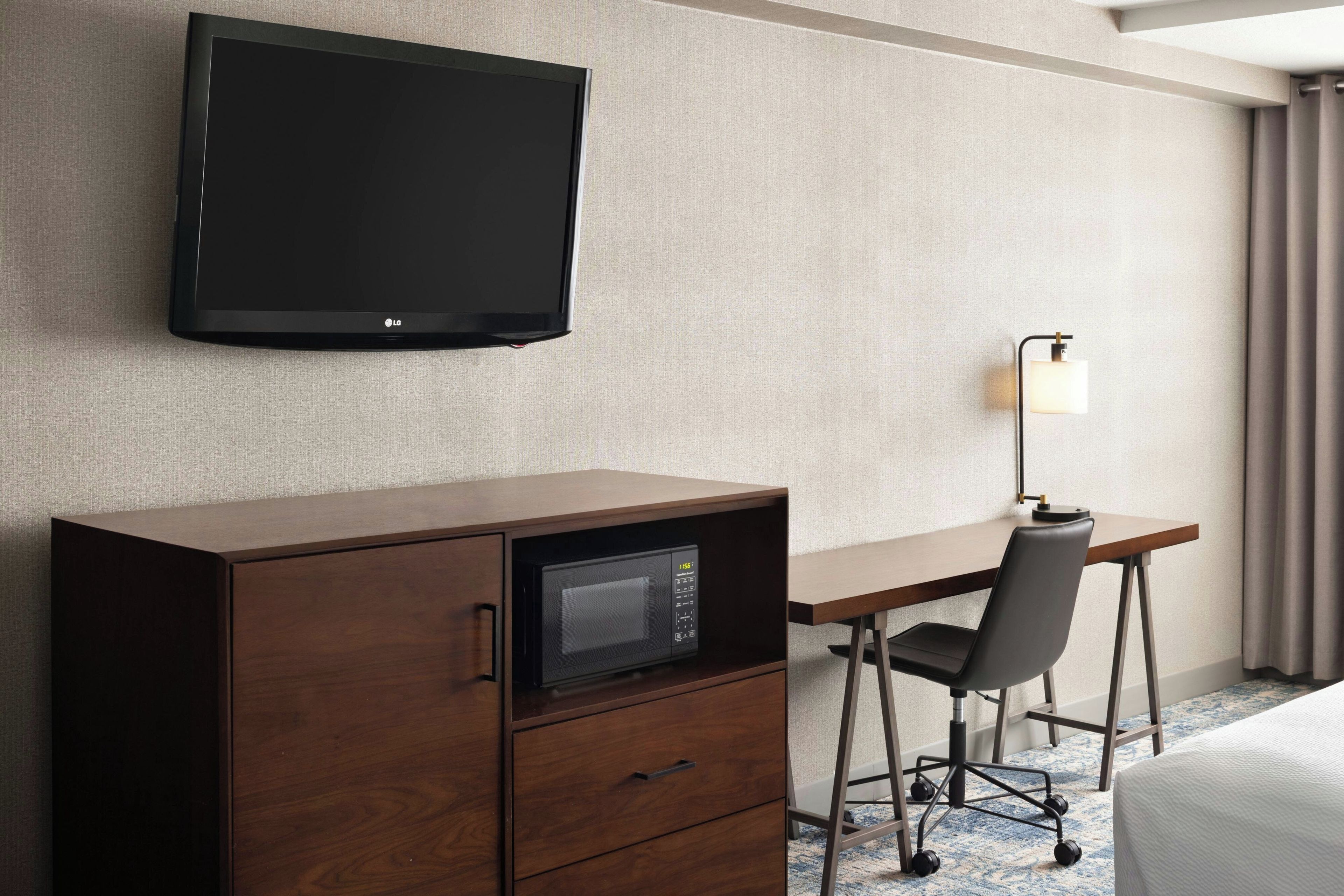 A hotel room with a flat-screen TV mounted on the wall, a wooden desk with a microwave, a chair, and a desk lamp.